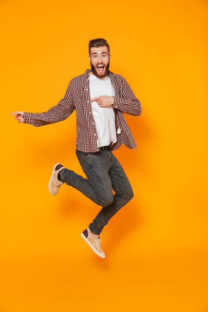 Full length portrait of a cheerful young man wearing casual clothes isolated over yellow background, pointing away at copy space, jumping - Foto, Imagen