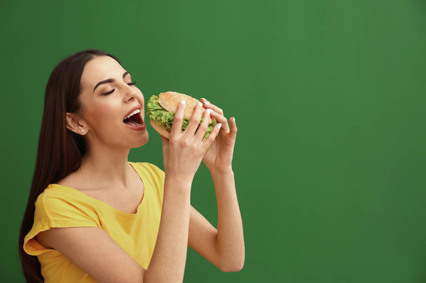 Young woman eating tasty burger on color background. Space for text - 写真・画像