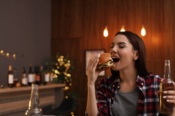 Young woman with beer eating tasty burger in cafe - Fotoğraf, Görsel
