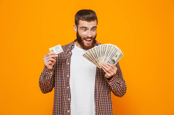 Portrait of a cheerful young man wearing casual clothes standing isolated over yellow background, holding money banknotes, showing plastic credit card - Foto, immagini