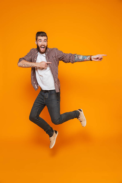 Full length portrait of a cheerful young man wearing casual clothes isolated over yellow background, jumping, pointing away - Foto, Bild