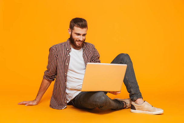 Arckép vidám fiatal férfi alkalmi ruhában, elszigetelt ül sárga háttérrel rendelkező laptop számítógép - Fotó, kép