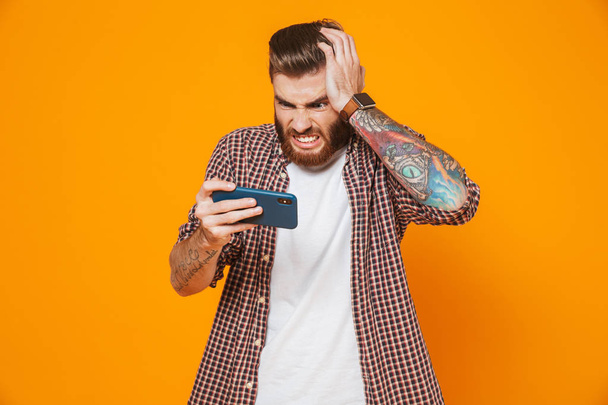 Portrait of an angry young man wearing casual clothes standing isolated over yellow background, playing games on mobile phone - Zdjęcie, obraz