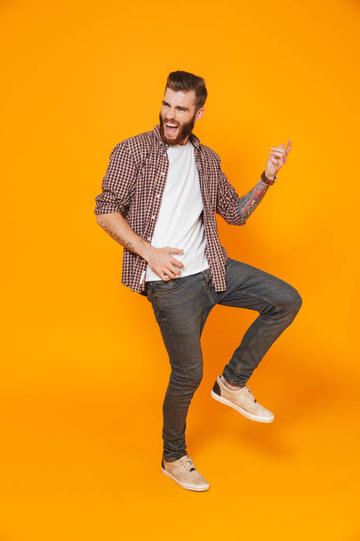 Full length portrait of a cheerful young man wearing casual clothes isolated over yellow background, playing on a pretend guitar - Фото, изображение