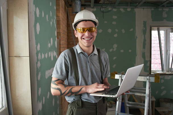 Room repair. Builder in helmet and glasses stands with a laptop in his hands on the background of the construction - Photo, Image