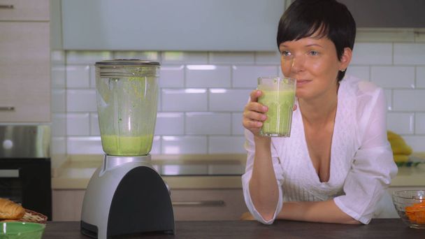 Volwassen vrouw drinken groene groenten smoothie in de keuken. Vrouw met korte kapsel dragen witte nachtkleding relish SAP in appartement in de ochtend. Gelukkig lachend brunette thuis. - Foto, afbeelding