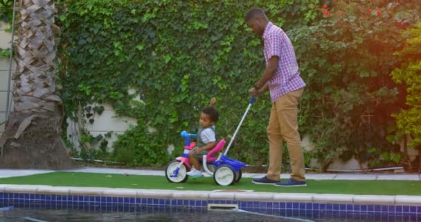 Side view of young black father and little son playing in back yard of their home. Father pushing tricycle 4k - Materiaali, video