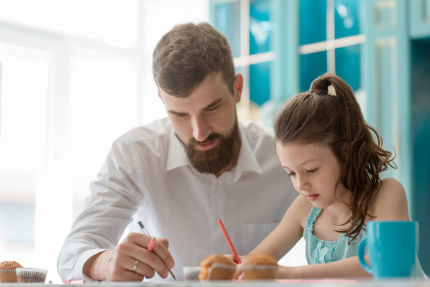 Father and his daughter spend quality time together, drawing a picture, drinking tea and eating cookies. Awesome parent concept. - Фото, изображение