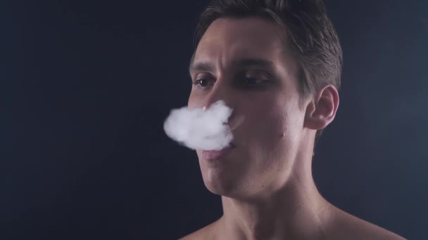 Vaper hombre guapo de cerca exhalando grandes nubes de humo con vapor de cigarrillo electrónico sobre fondo negro en cámara lenta
 - Imágenes, Vídeo