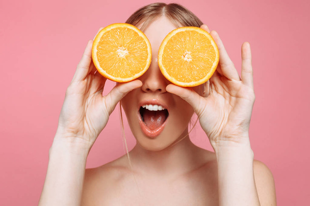 Portrait of a cheerful feminine girl, with natural clear skin, a girl with orange slices, covering her eyes, isolated on a pink background. Skin care concept, natural beauty - Photo, Image