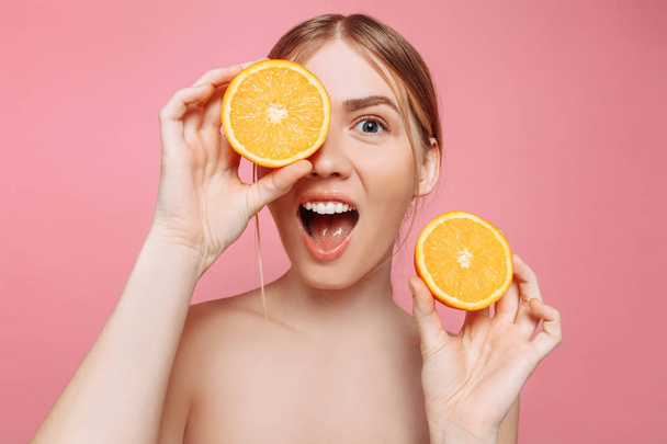 Portrait of a cheerful shocked feminine girl, with natural clear skin, girl with orange slices, isolated on a pink background. Skin care concept, natural beauty - Photo, image