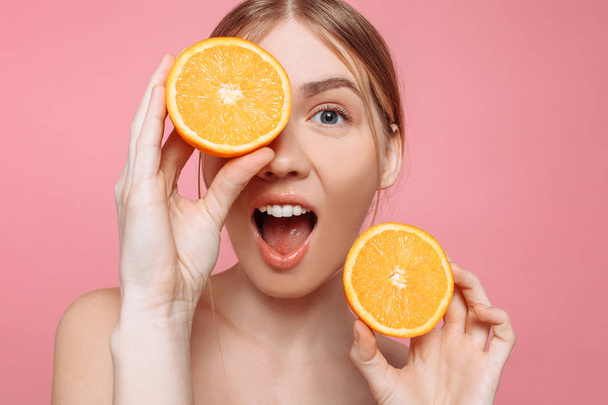 Portrait of a cheerful shocked feminine girl, with natural clear skin, girl with orange slices, isolated on a pink background. Skin care concept, natural beauty - Фото, изображение