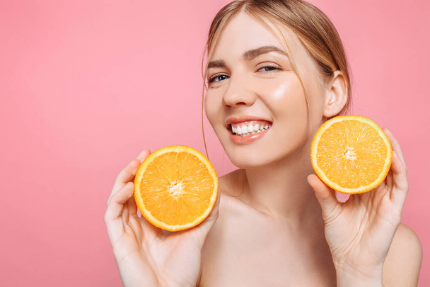 Portrait of a cheerful feminine girl, natural clear skin, girl with two orange slices, isolated on a pink background. Skin care concept, natural beauty - Photo, image