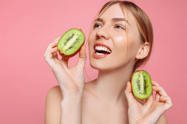 Portrait of a cheerful feminine girl, natural clear skin, a girl with two pieces of kiwi, isolated on a pink background. Skin care concept, natural beauty - Fotoğraf, Görsel