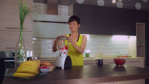 Καυκάσιος μελαχρινή με κοντό κούρεμα στο σπίτι μετά την προπόνηση, προετοιμάζοντας smoothie με φρούτα. Γυναίκα να στέκεται στην κουζίνα στο τραπέζι μπολ με φράουλα. Υγιεινός τρόπος ζωής. - Φωτογραφία, εικόνα