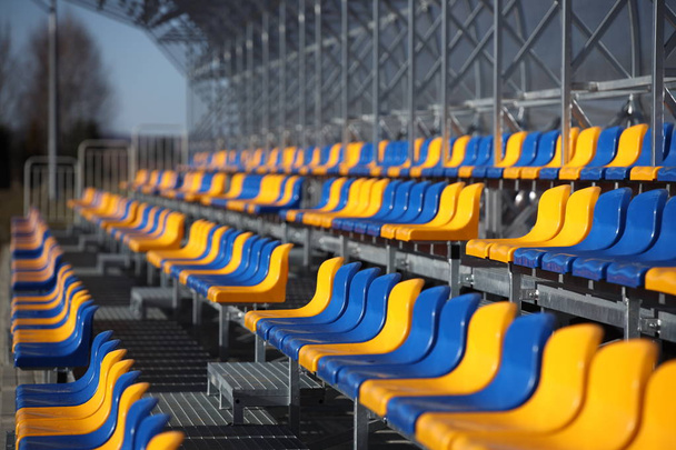 Nuevos stands en el campo de fútbol de construcción ligera de metal con asientos de plástico en azul y amarillo. Lugares para aficionados en el estadio. Ir a la audiencia para competiciones deportivas. Refugio de la carrera
. - Foto, Imagen