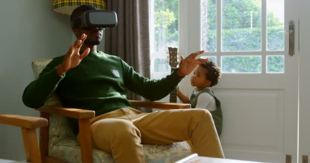 Front view of young black father using virtual reality headset in a comfortable home. Son playing with guitar 4k - Záběry, video
