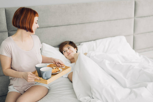 attractive girl bringing breakfast to her sister lying on the bed - Fotó, kép