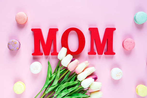 Woord moeder van rode letters en tedere tulpen, macaroons op roze achtergrond. Moeders dag decoratie concept. Bovenaanzicht, platte lay met kopieerruimte - Foto, afbeelding