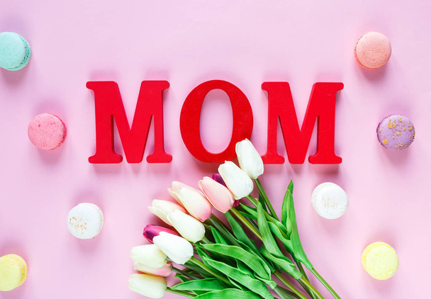 Wortmama aus roten Buchstaben und zarten Tulpen, Makronen auf rosa Hintergrund. Muttertagsdekoration. Draufsicht, flache Lage mit Kopierraum - Foto, Bild