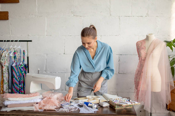 Young female tailor sits at workplace and chooses color of threads in atelier - Foto, Bild