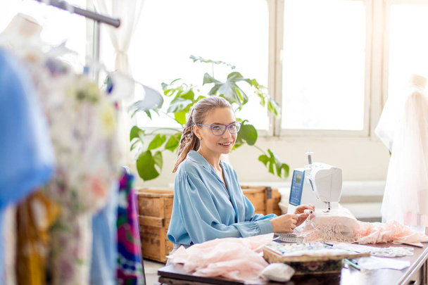 Young female tailor sits at workplace and chooses color of threads in atelier - Valokuva, kuva
