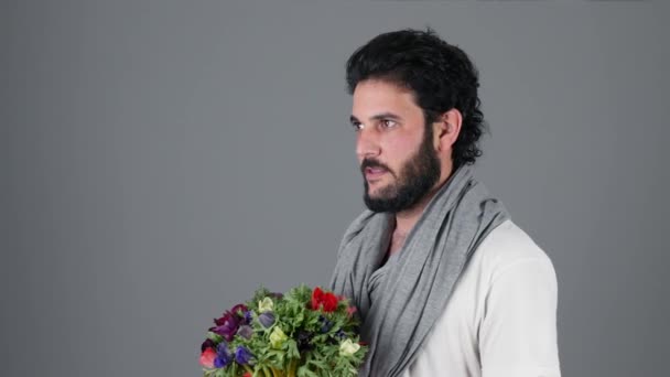 Handsome young man over grey wall holding flowers bouquet and with showing teeth - Video