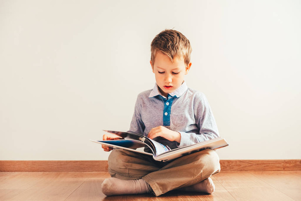 Chłopiec siedzi na podłodze z czytając książkę na kolanach, na białym tle. - Zdjęcie, obraz