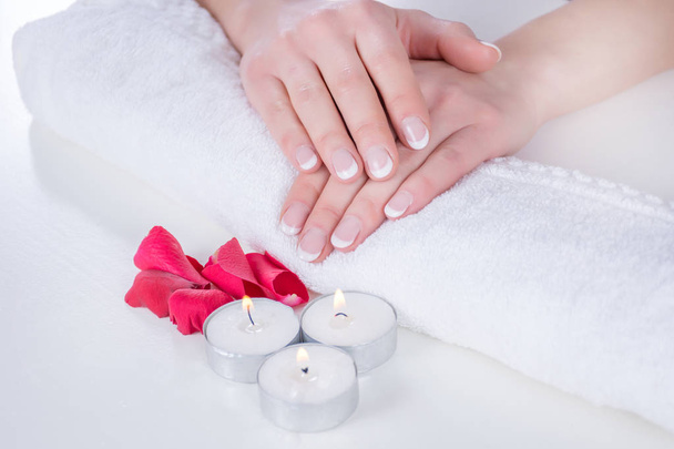 Woman hands with french manicure modern style on towel with red rose petals and candle in beauty salon. Manicure and Beauty concept. Close up, selective focus - Photo, Image