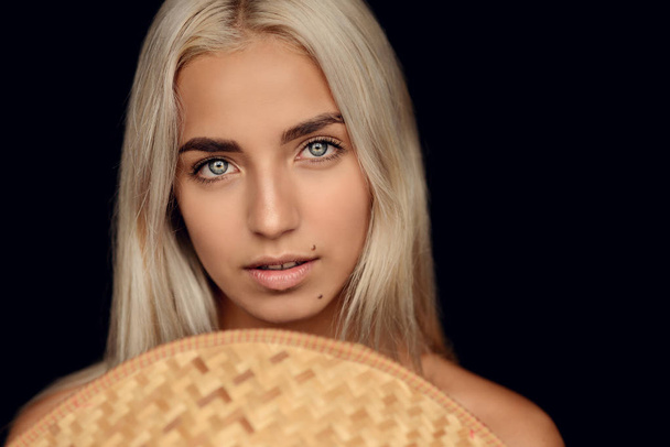 fashion photo of beautiful woman with blonde hair and expressive eyes posing with asian style wicker hat on the black background in studio - Photo, image