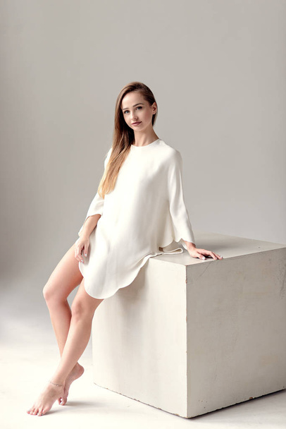 fashion photo of beautiful woman with brunette hair and long attractive legs in white elegance dress posing about big cube on white background in studio. - 写真・画像