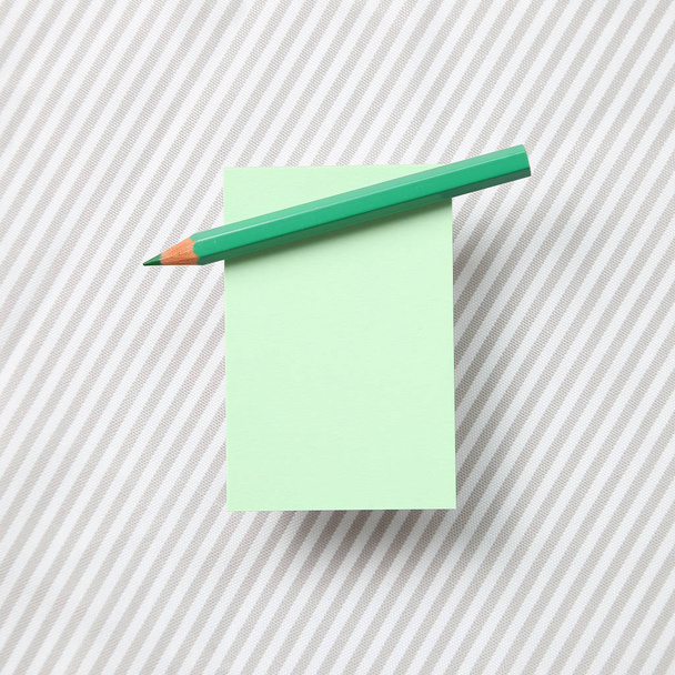 Green memo note pad and green colored pencil on striped pattern fabric background - Photo, Image