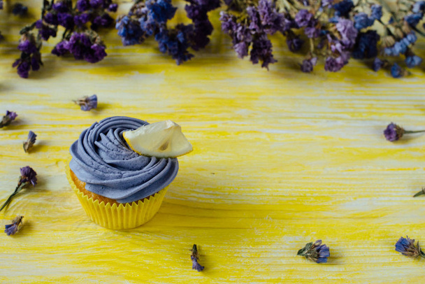 Close up of single cupcake decorated with coloured whipped cream and a piece of lemon on a yellow wooden background in shabby chic style and dried flowers at the background and petals of flowers at the front.Text space - Photo, Image