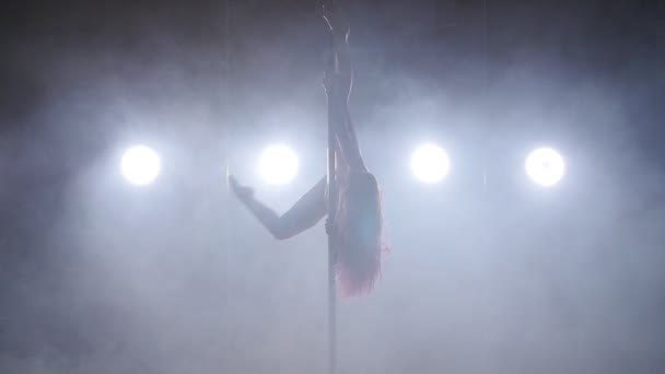 Concept van dans en acrobatiek. Jonge slanke vrouw paaldansen in donkere interieur met achtergrondverlichting en rook - Video