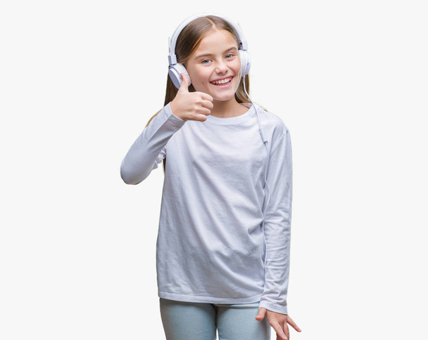 Mooi meisje koptelefoon luisteren naar muziek op geïsoleerde achtergrond doen gelukkig duimschroef opwaarts gebaar met de hand dragen. Goedkeuring van expressie kijken naar de camera met tonen van succes. - Foto, afbeelding