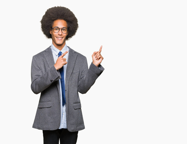 Joven hombre de negocios afroamericano con cabello afro con gafas sonriendo y mirando a la cámara apuntando con dos manos y dedos hacia un lado
. - Foto, imagen