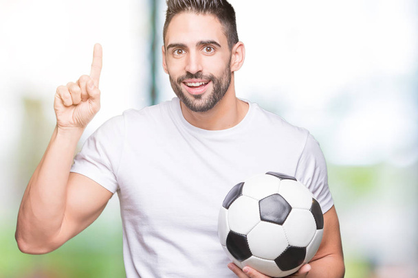 Молодой человек держит футбольный мяч на изолированном фоне, удивленный идеей или вопросом, указывая пальцем со счастливым лицом, номер один
 - Фото, изображение