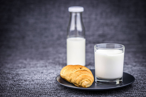 Croissant em uma chapa com um copo de leite e meia garrafa de leite em um fundo cinza
 - Foto, Imagem