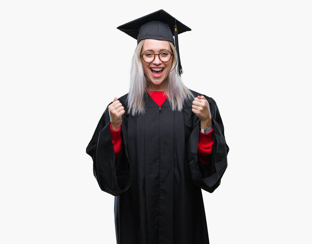 Young blonde woman wearing graduate uniform over isolated background celebrating surprised and amazed for success with arms raised and open eyes. Winner concept. - Фото, изображение