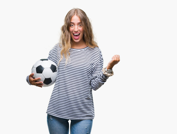 Hermosa joven rubia sosteniendo pelota de fútbol sobre fondo aislado gritando orgulloso y celebrando la victoria y el éxito muy emocionado, animando emoción
 - Foto, Imagen