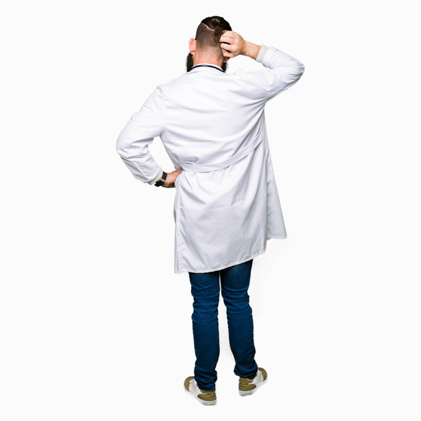 Young blond doctor man with beard wearing medical coat Backwards thinking about doubt with hand on head - Foto, Bild
