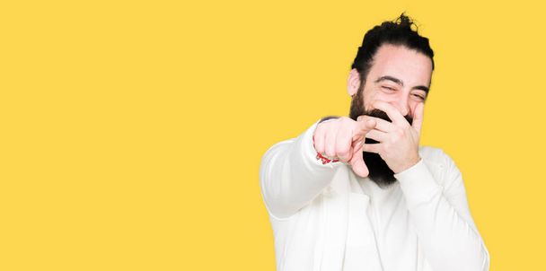 Young hipster man with long hair and beard wearing sporty sweatshirt Laughing of you, pointing to the camera with finger hand over mouth, shame expression - 写真・画像
