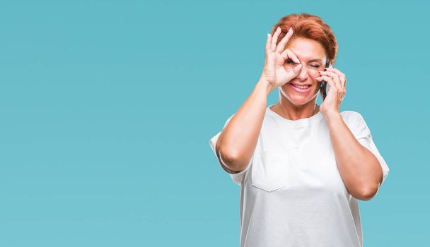 Atrractive senior caucasian redhead woman talking on smartphone over isolated background with happy face smiling doing ok sign with hand on eye looking through fingers - Fotografie, Obrázek