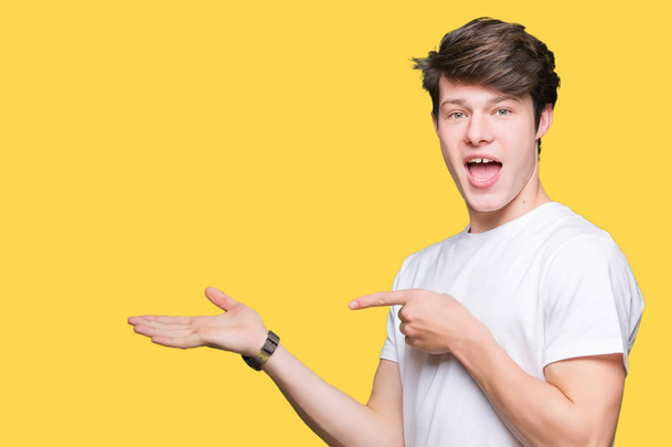 Young handsome man wearing casual white t-shirt over isolated background amazed and smiling to the camera while presenting with hand and pointing with finger. - Photo, Image