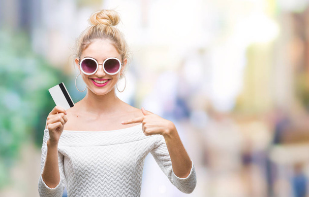 Young beautiful blonde woman holding credit card over isolated background with surprise face pointing finger to himself - Fotoğraf, Görsel