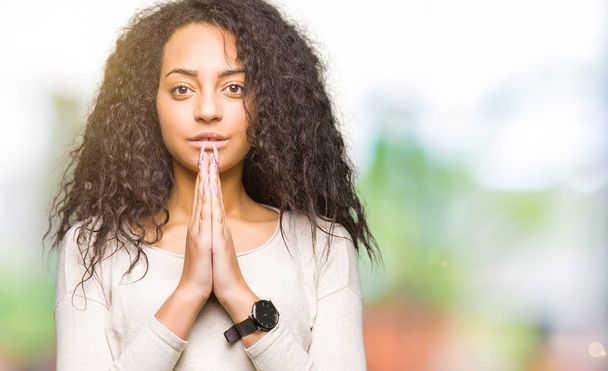 Young beautiful girl with curly hair wearing casual sweater praying with hands together asking for forgiveness smiling confident. - Photo, Image