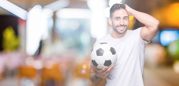 Jonge man houdt van voetbal voetbal over geïsoleerde achtergrond benadrukt met de hand op het hoofd, geschokt met schaamte en verrassing gezicht, boos en gefrustreerd. Angst en boos voor fout. - Foto, afbeelding