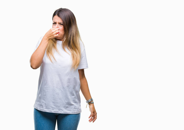 Young beautiful woman casual white t-shirt over isolated background smelling something stinky and disgusting, intolerable smell, holding breath with fingers on nose. Bad smells concept. - Фото, зображення