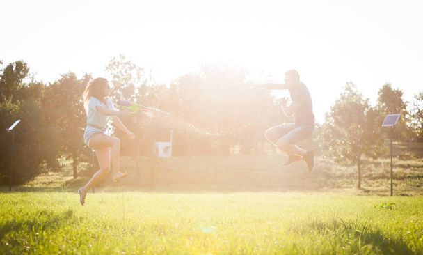 Ένα παιχνιδιάρικο ζευγάρι κυνηγώντας ο ένας τον άλλον και να παίζει με νεροπίστολα σε ένα λιβάδι ar ηλιοβασίλεμα. - Φωτογραφία, εικόνα