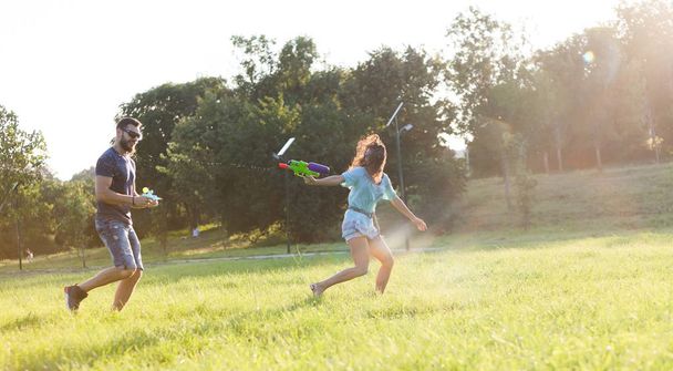 A playful young couple chasing each other and playing with water guns in a meadow ar sunset. - Photo, Image
